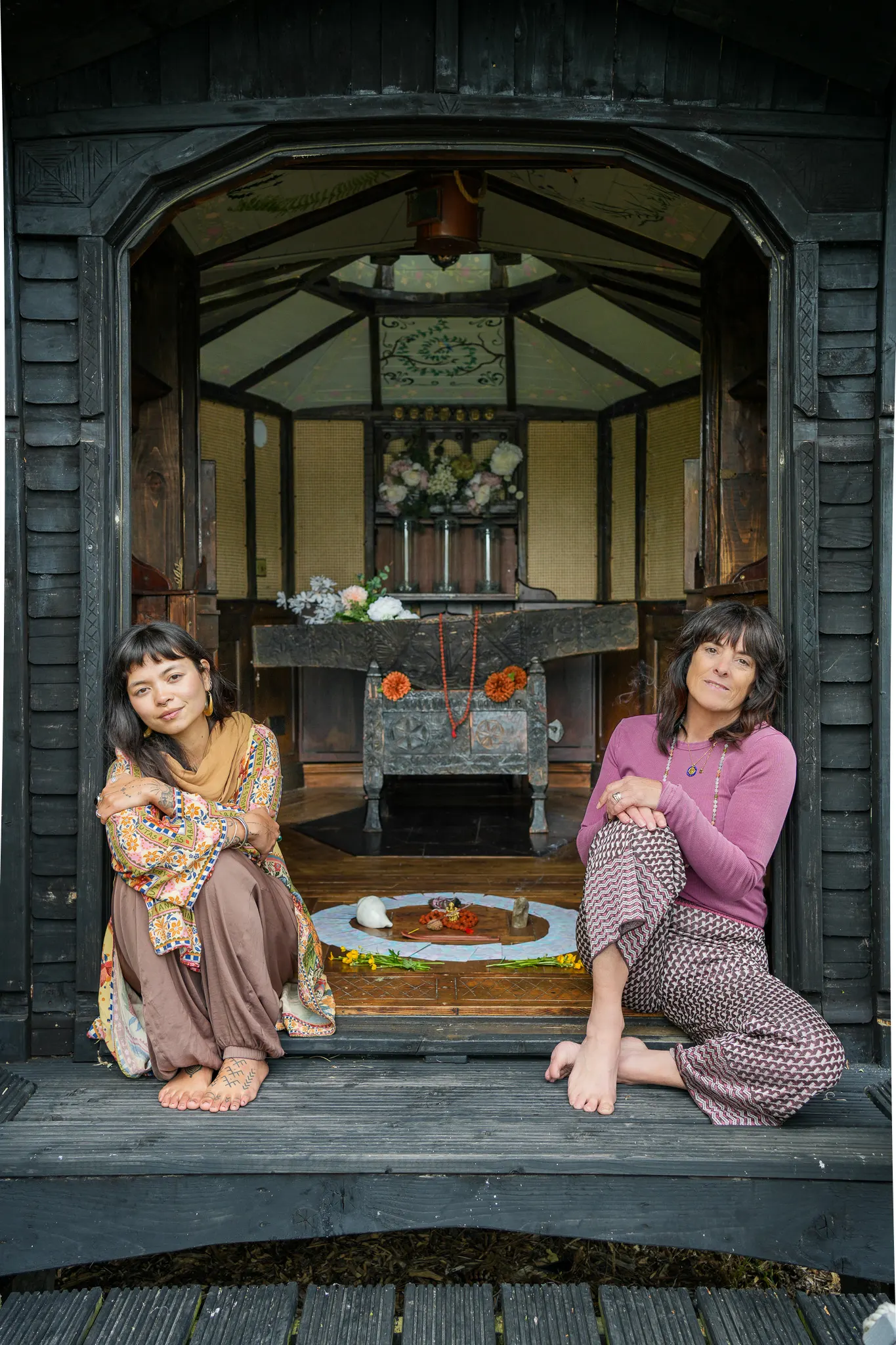 Catherine and Dani sitting barefoot in the doorway of an ornate wooden summer house, an altar behind them