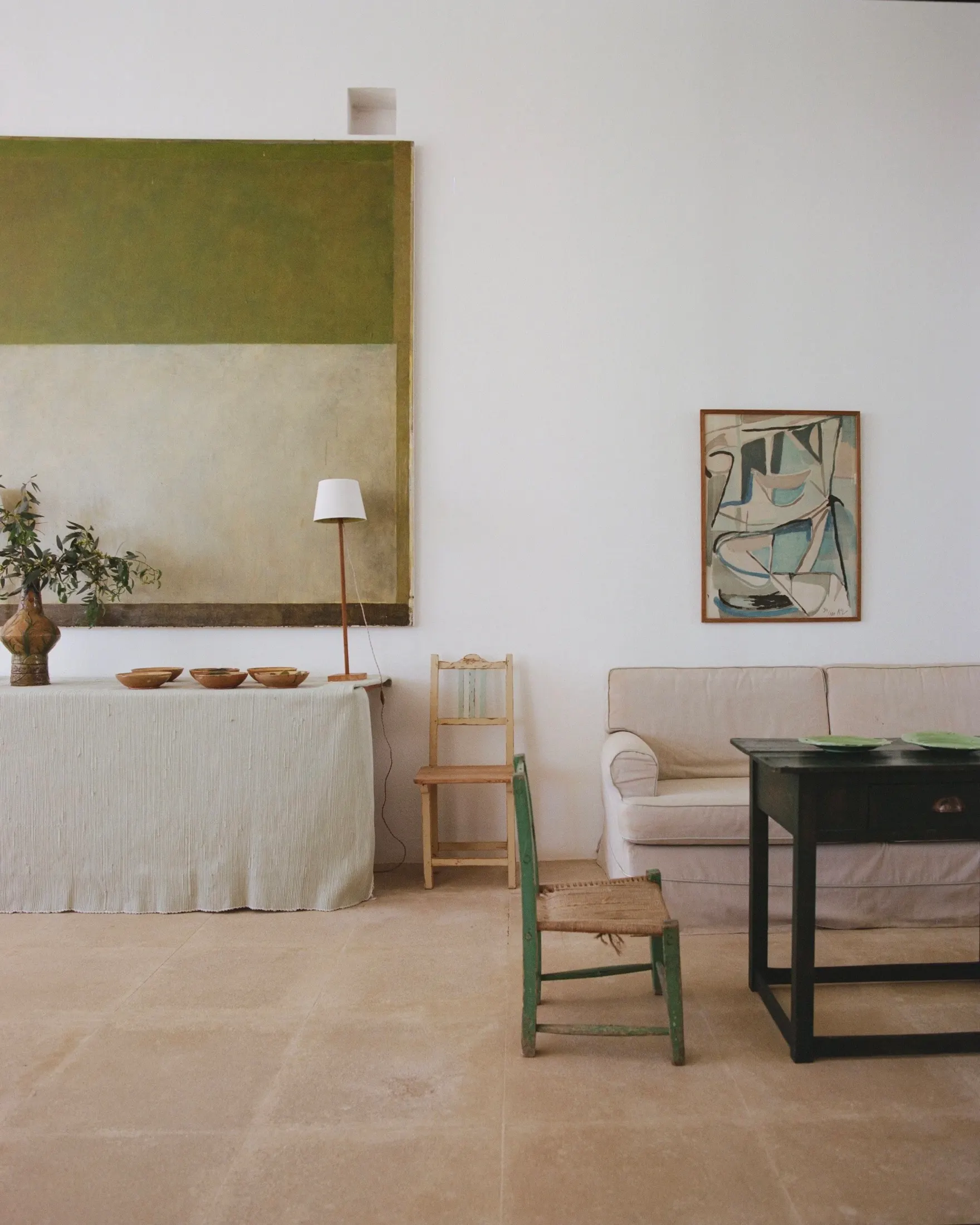 A calm white sitting room with a green painting and simple wooden chairs